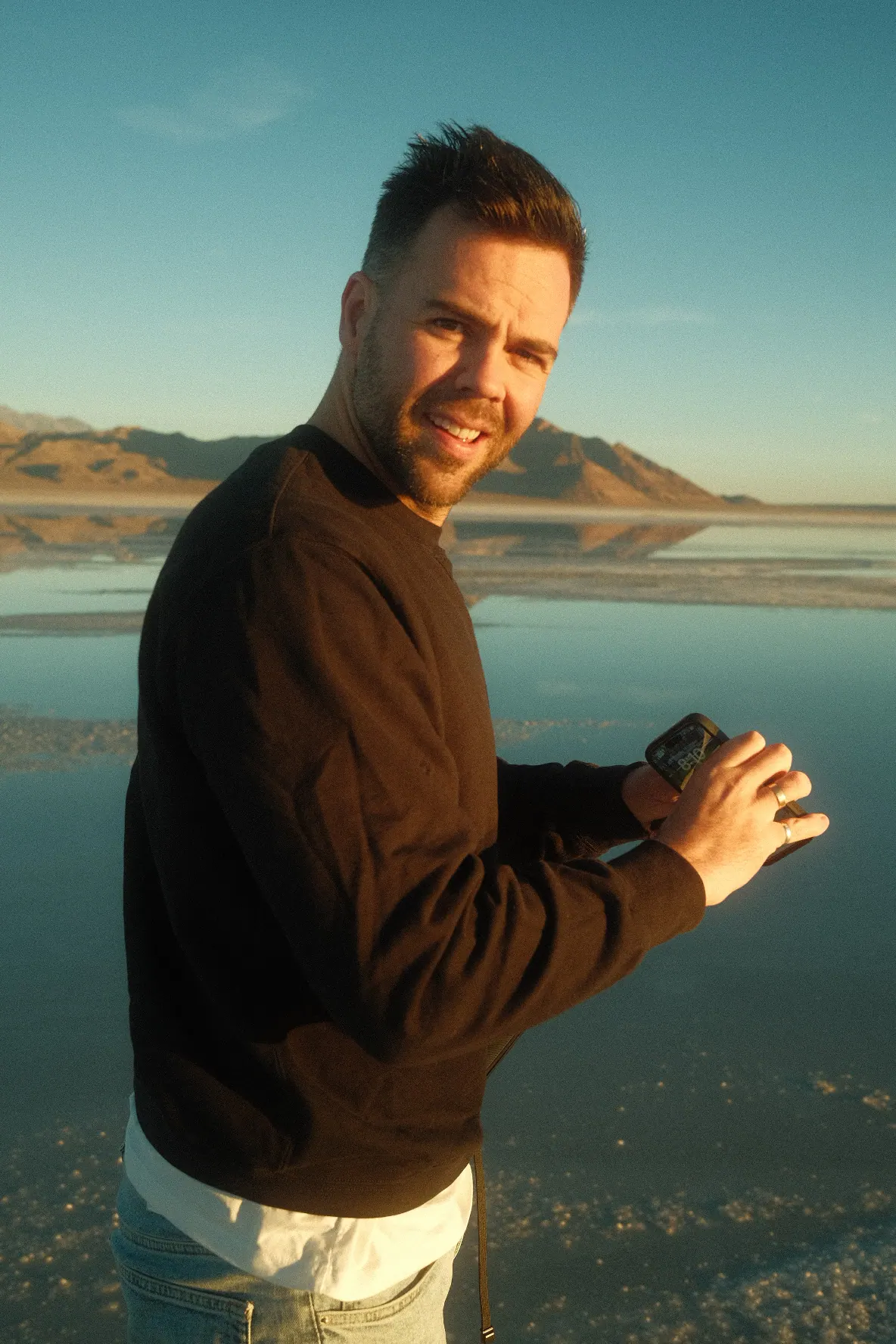 Tommy Jackett in a dark jumper smiling at the camera, standing in shallow water on the Bonneville Salt Flats at sunset.